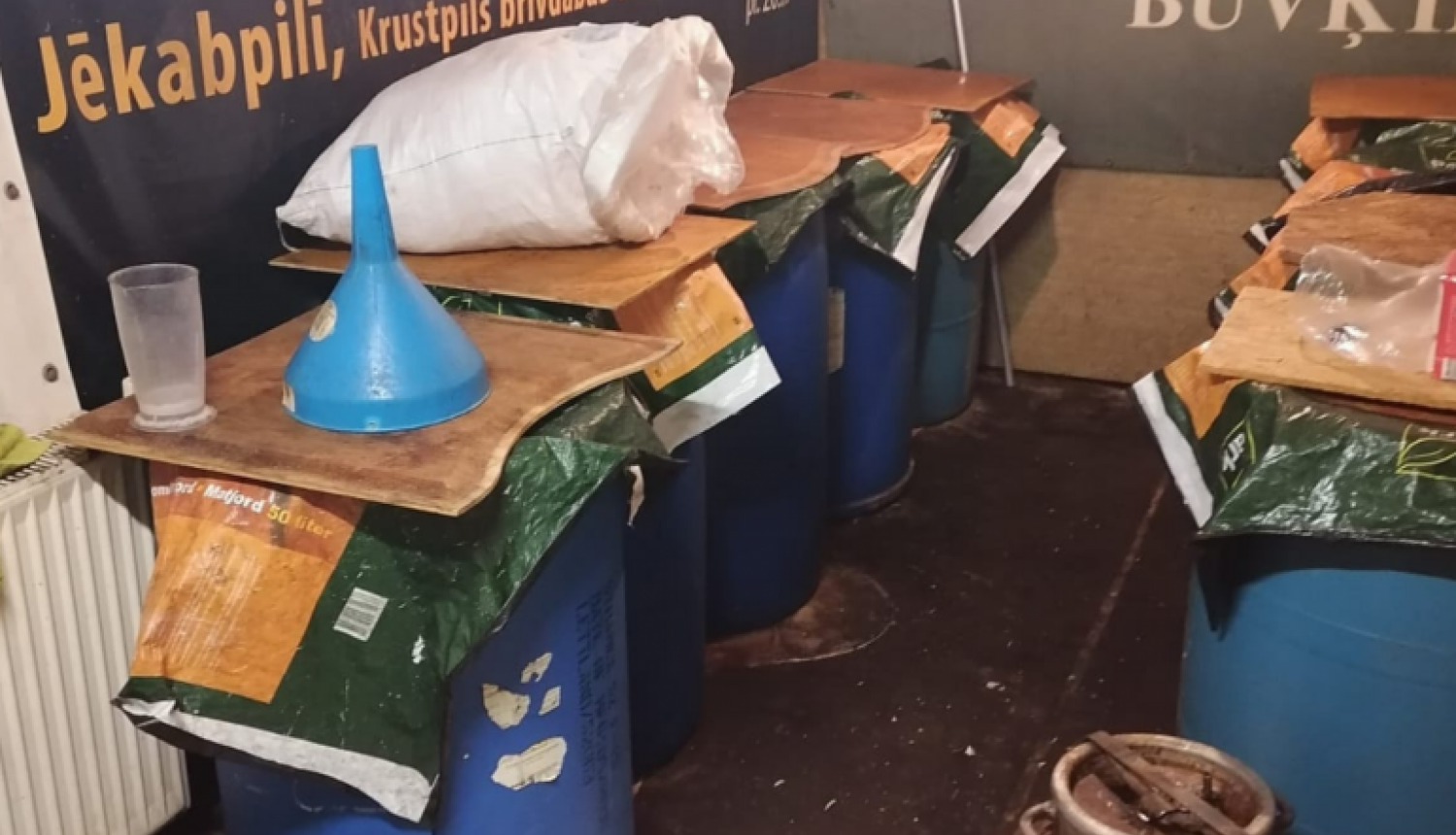 Zilas plastmasas mucas, pārsegtas ar finiera gabaliem, priekšplānā alumīnija kanna, fonā pie sienas operas izrādes "Sikspārnis" platāts un uzņēmuma "Vincents" reklāma  