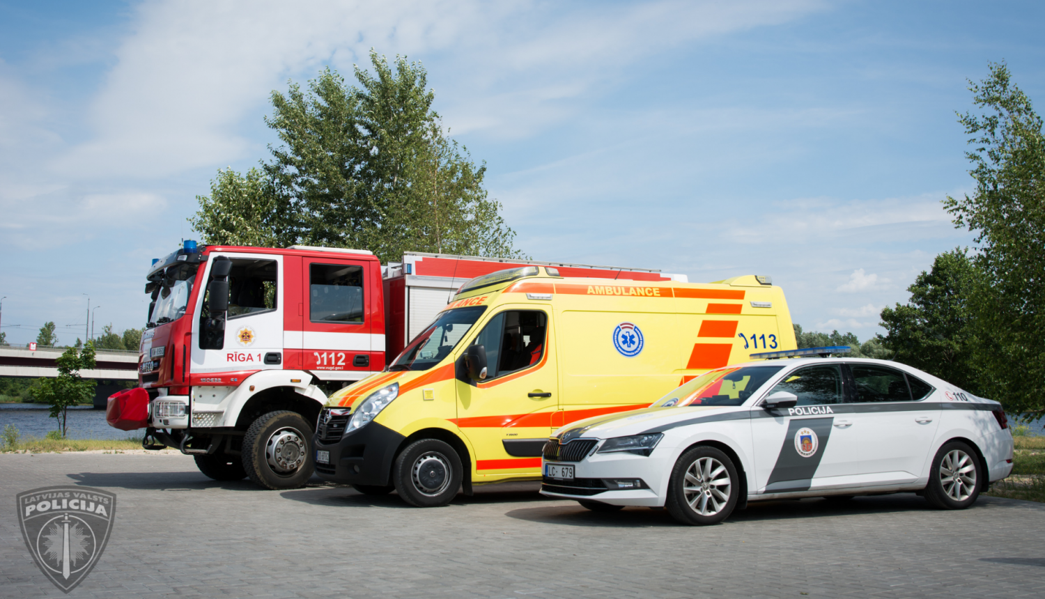 Pie upes novietoti stāvēšanai cits citam blakus operatīvie transportlīdzekļi, priekšplānā policijas mašīna, pa vidu neatliekamās medicīniskās palīdzības dienesta mašīna, tālāk valsts ugunsdzēsības un glābšanas dienesta mašīna