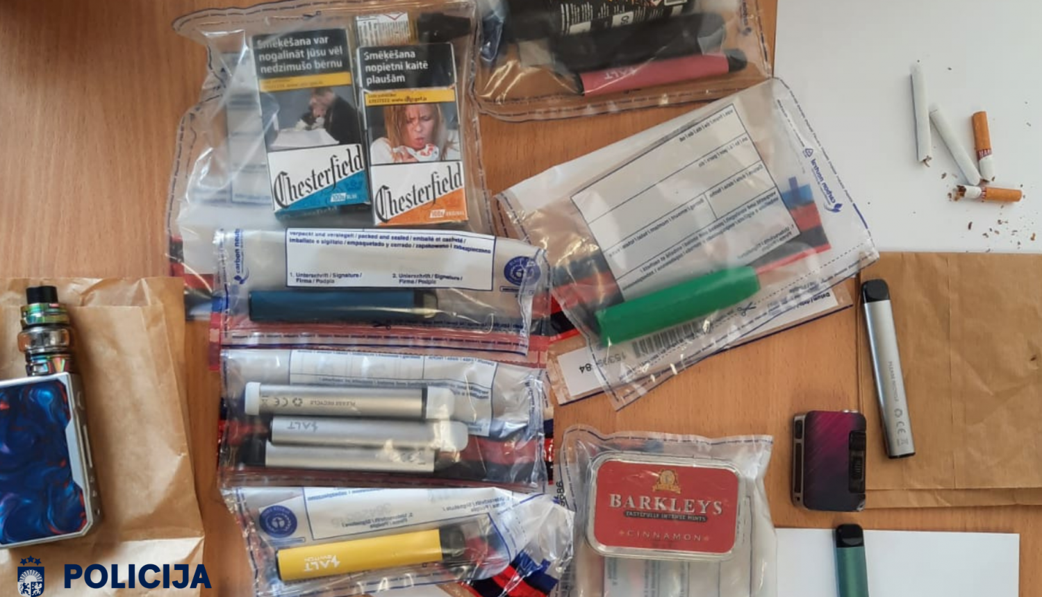 Uz galda dažādas elektroniskās smēķēšanas ierīces, cigarešu paciņas, uzpildes šķidruma tvertnes un cigaretes