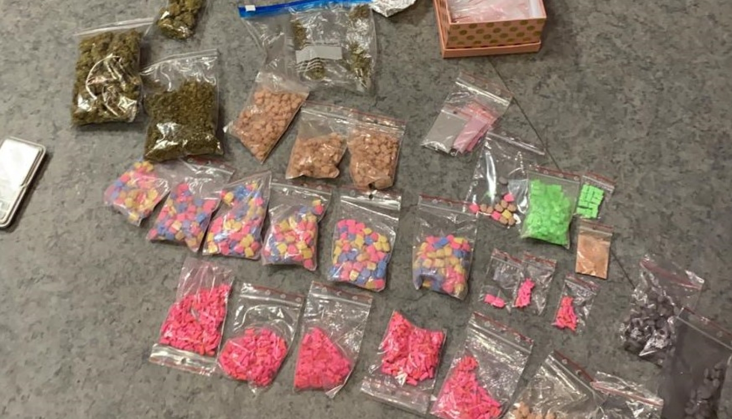 Uz grīdas nolikti vairāki caurspīdīgi plastmasas maisiņi ar dažādu krāsu tabletēm - narkotikām, marihuānu, folijā ietītas markas, pelēks mobilais tālrunis