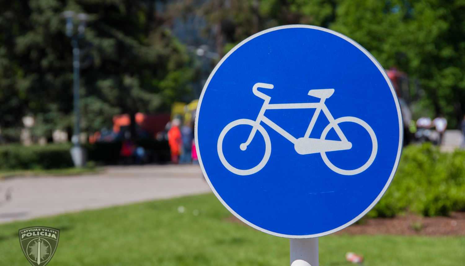 Ceļa zīme, kas norāda riteņbraucēju ceļu, fonā redzams parks