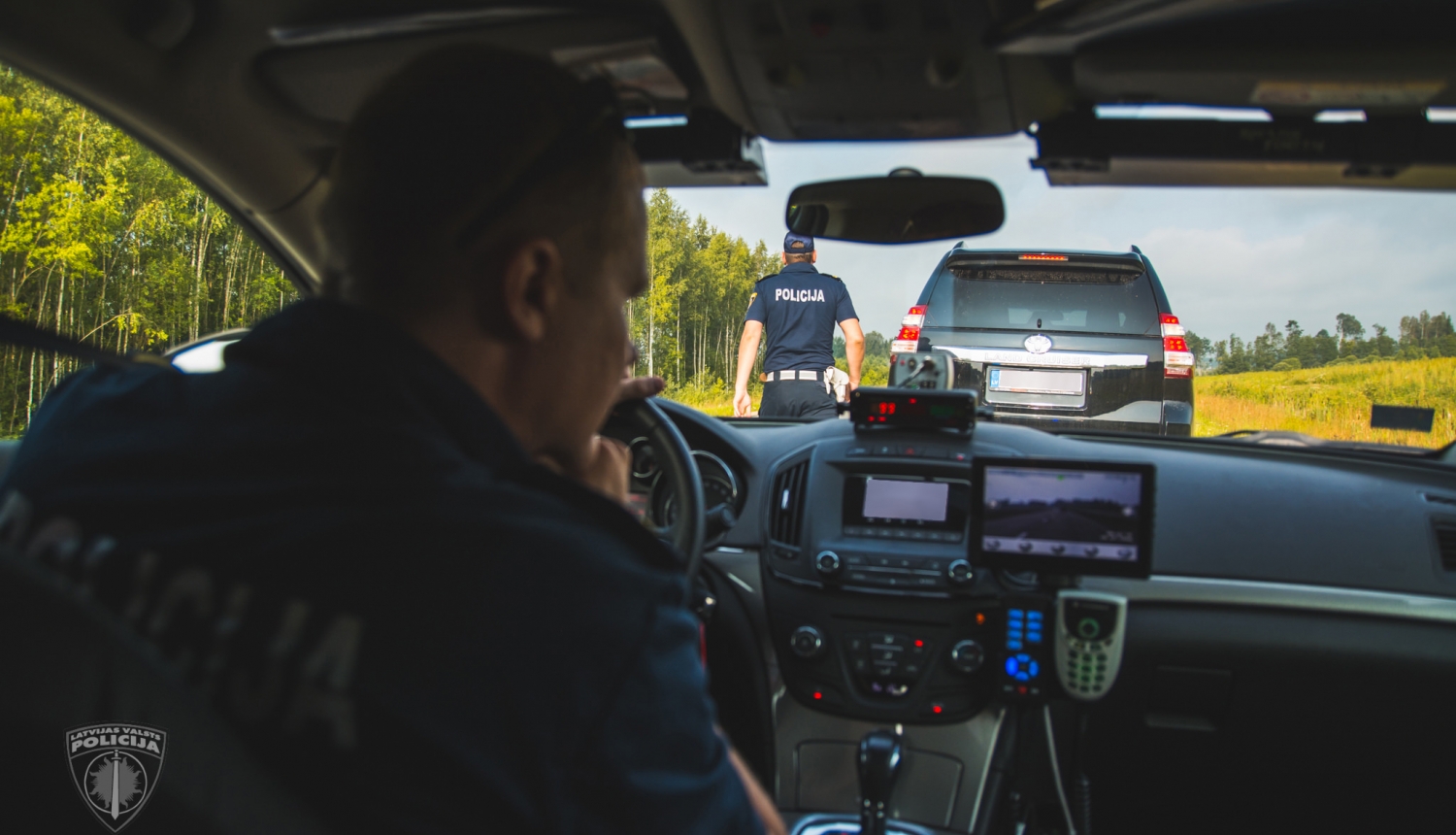 Skats policijas mašīnas iekšienē, pie stūres policists dienesta formā