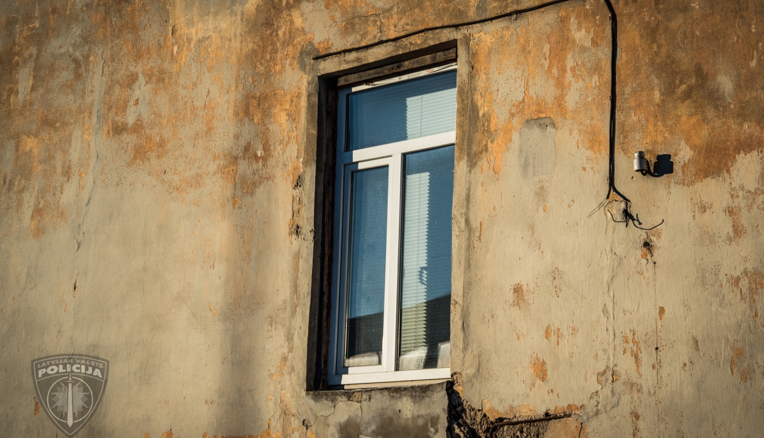 Mājas siena, viens logs ar horizontālajām žalūzijām, stūrī zem loga izdrupusi iedobe sienā