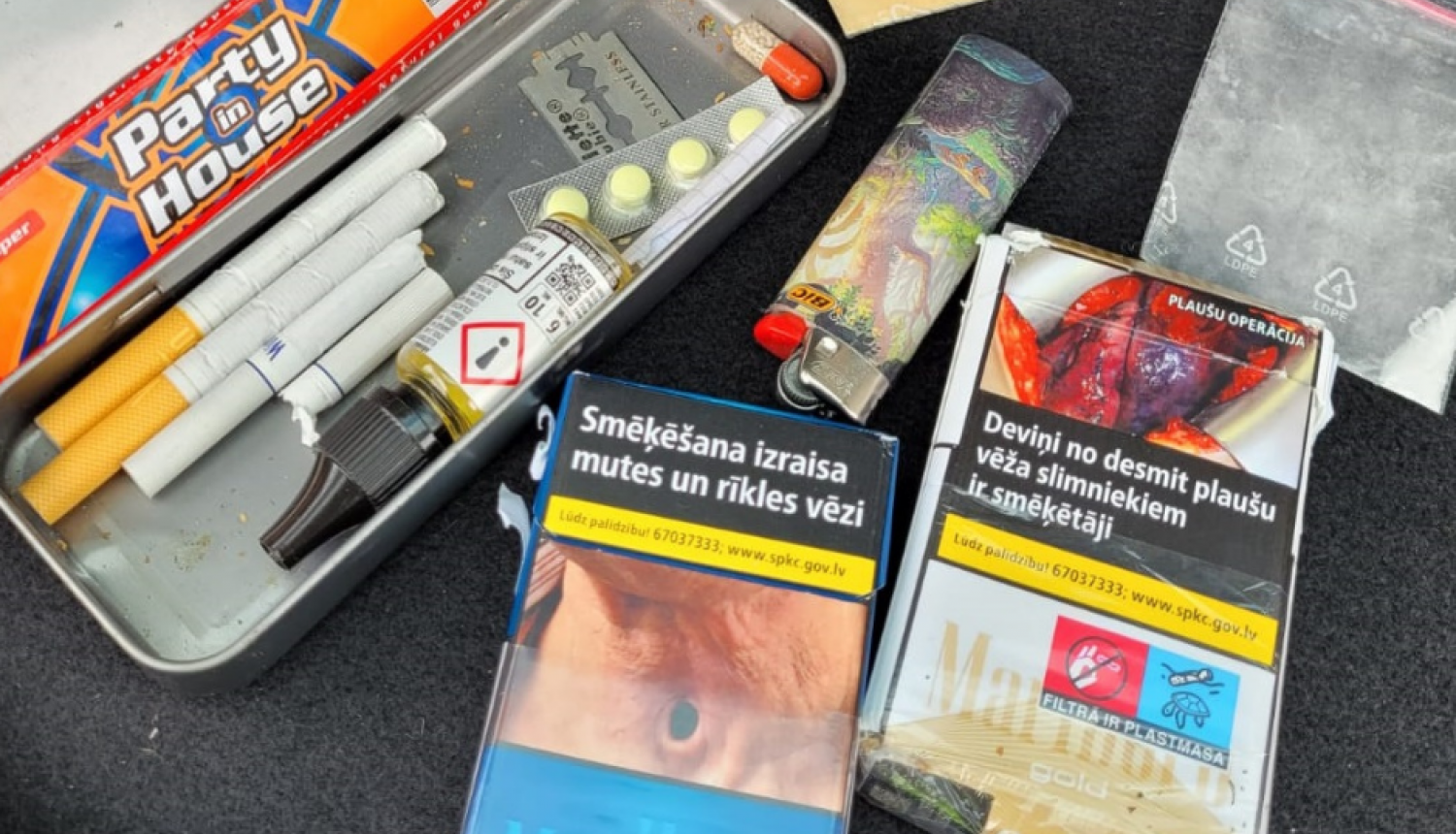 Metāla penālis, kurā atrodas cigaretes, tabletes, žilete, pie penāļa cigarešu paciņas, caurspīdīgi plastikāta maisiņi ar baltas un oranžas krāsas pulverīšiem