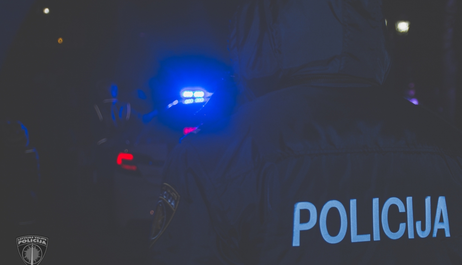Tumsā redzamas policijas dienesta automašīnas zilā bākuguns un uzraksts "POLICIJA" uz policista dienesta formas mugurpuses