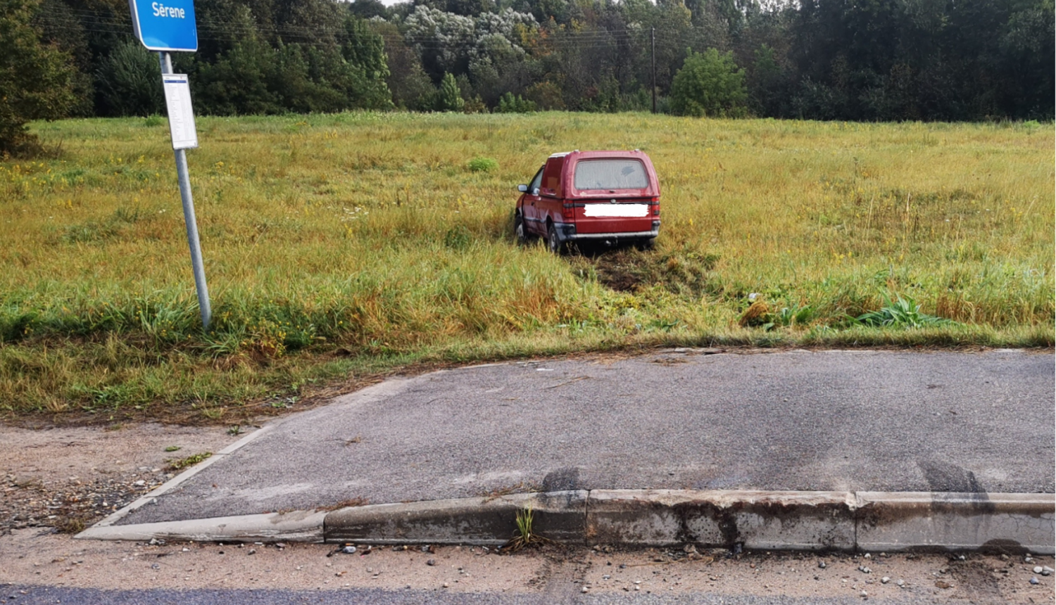 Sarkana automašīna iebraukusi pļavā, priekšplānā autobusa pietura