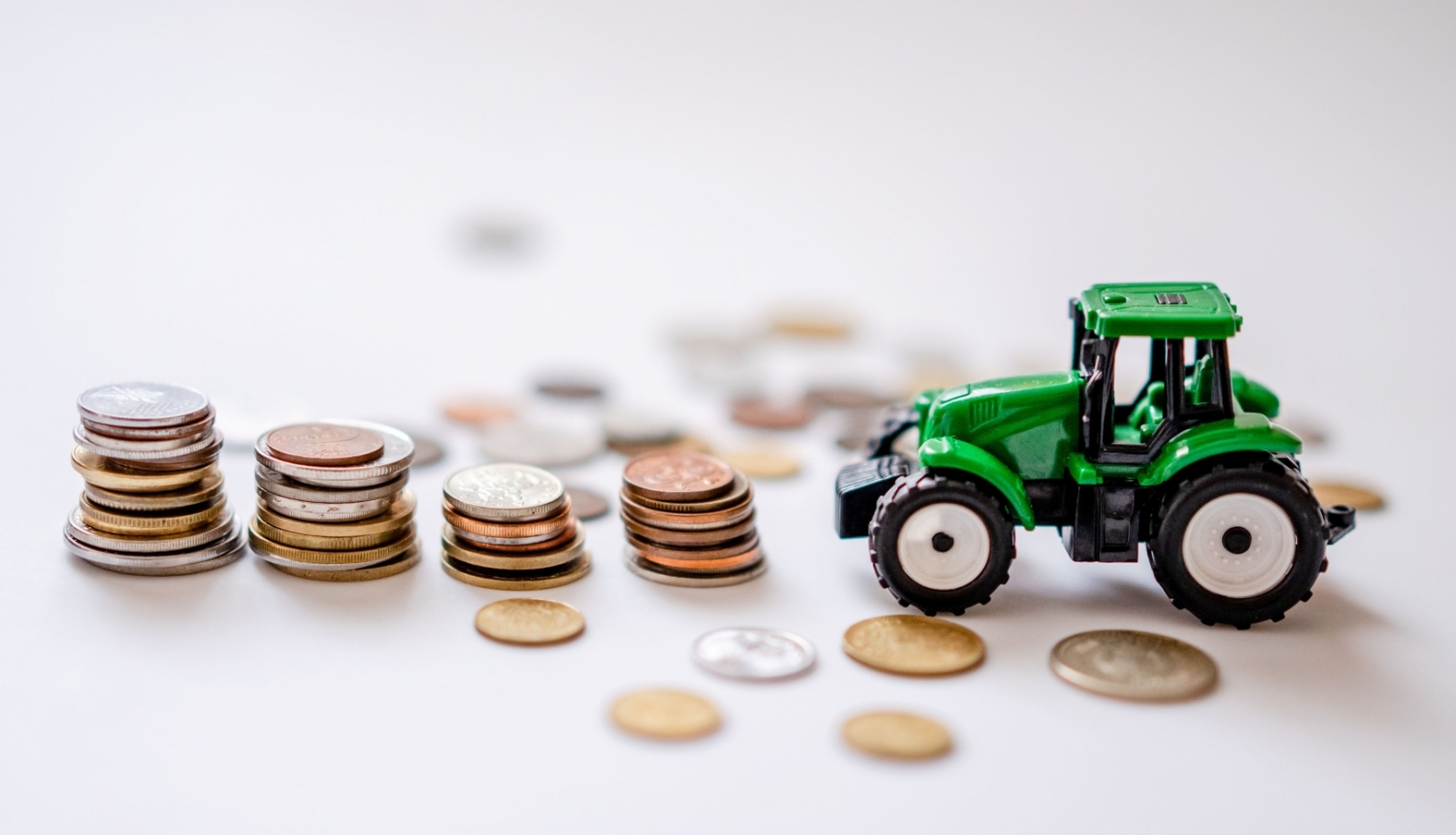 Zaļš rotaļu traktors, kuram priekšā četras monētu kaudzītes un atsevišķas monētas izbārstītas pa gaišu galda virsmu