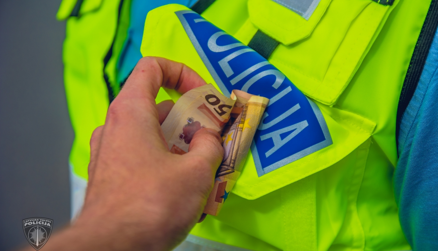 Plaukstā redzami 50 eiro, ko cenšas ielikt policista formas apģērba vestes priekšējā kabatā