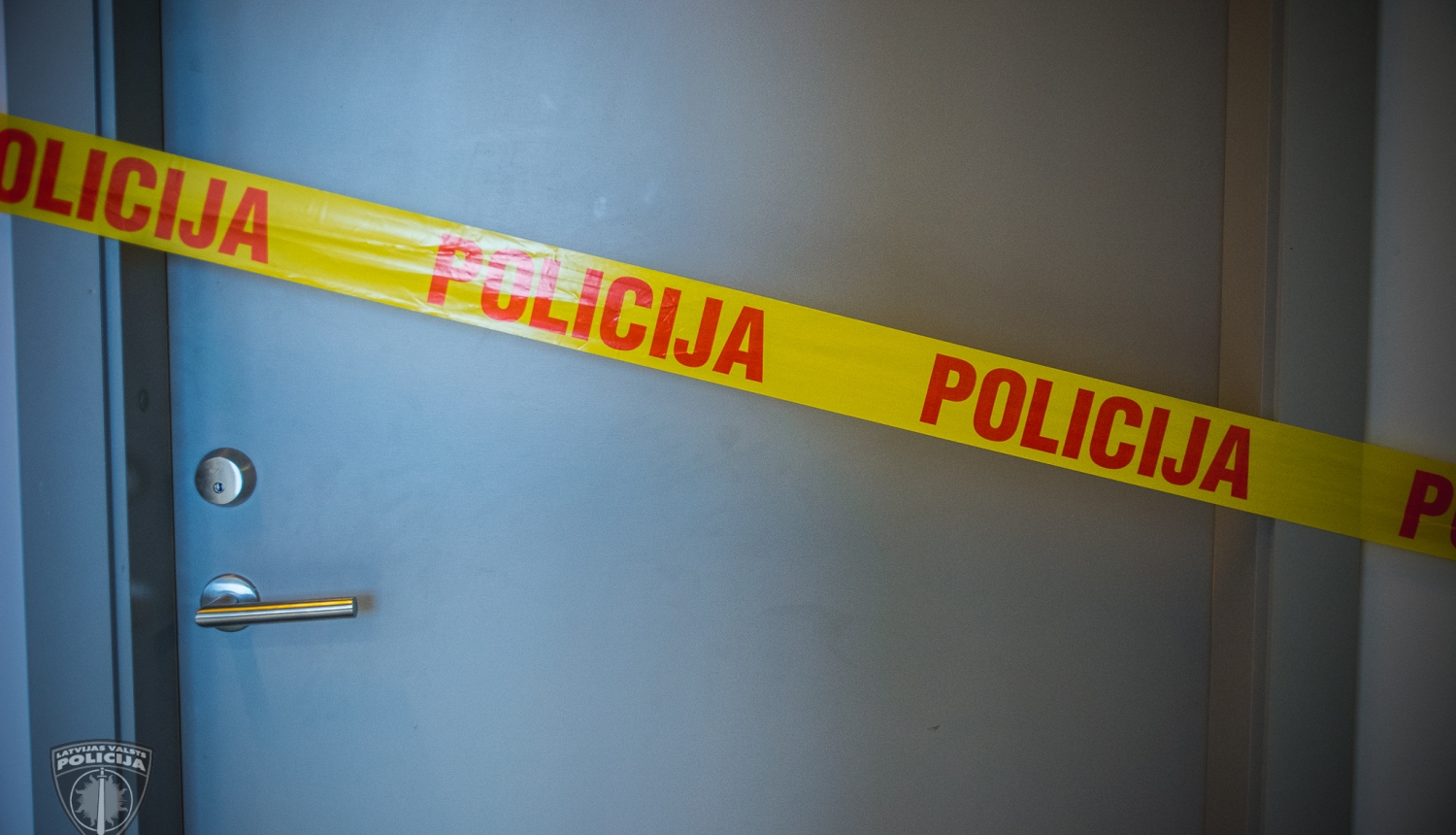 Pelēkas durvis, kurām priekšā novilkta dzeltena lenta, uz kuras ar sarkaniem burtiem uzrakstīts "Policija"